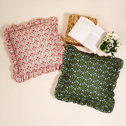 The Darling Effect Everbloom Forest Green Scalloped Square Throw Pillow rests on a light surface beside another pillow, a woven mat, an open book, and a small bouquet of white flowers for a cozy decorative touch.