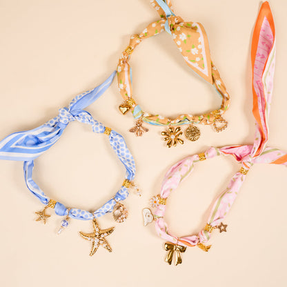 Three Evergreen Twist Make a Statement Scarf Necklaces by The Darling Effect—blue polka-dot with starfish and shell charms, floral tan with bee and daisy charms, and pink floral with gold bow—arranged on a beige background.