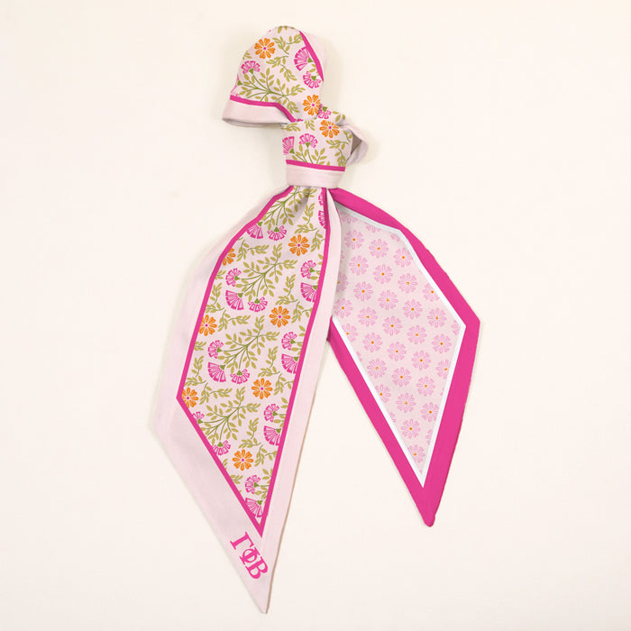 The Darling Effect’s Gamma Phi Beta Twirl & Tie Scarf Accessory features pink edges, floral patterns in pink, yellow, and green, plus embroidered TBP initials—styled here in a loose knot against a plain background.