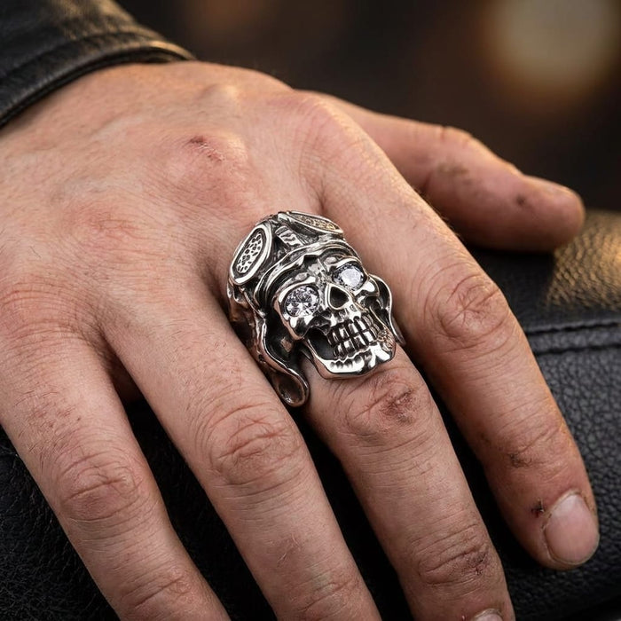 Close-up lifestyle shot of a man's rugged hand wearing a chunky silver German pilot skull ring featuring clear gemstone eyes and a sculpted aviator helmet, resting on a textured black leather surface.