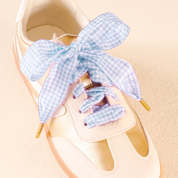 A white and gold sneaker features The Darling Effect's Gingham Light Blue Step It Up Shoelaces, tied in a large bow on top.