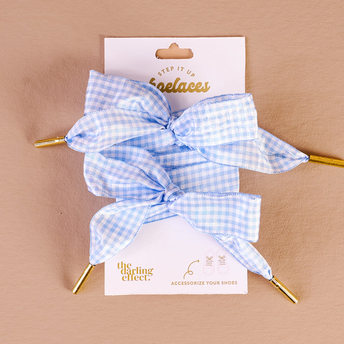 Two Gingham Light Blue Step It Up Shoelaces from The Darling Effect, each with gold aglets, are tied in bows and displayed on a white card. These durable laces add a stylish touch to your favorite sneakers.