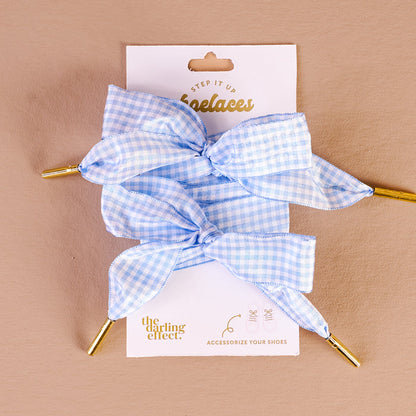 Two Gingham Light Blue Step It Up Shoelaces from The Darling Effect, each with gold aglets, are tied in bows and displayed on a white card. These durable laces add a stylish touch to your favorite sneakers.