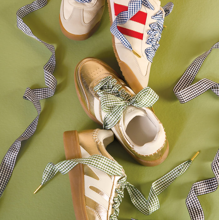 Three sneakers with gum soles on a green background, each laced with The Darling Effect's Gingham Light Blue Step It Up Shoelaces. One sneaker has red stripes, while the others feature beige and white detailing.