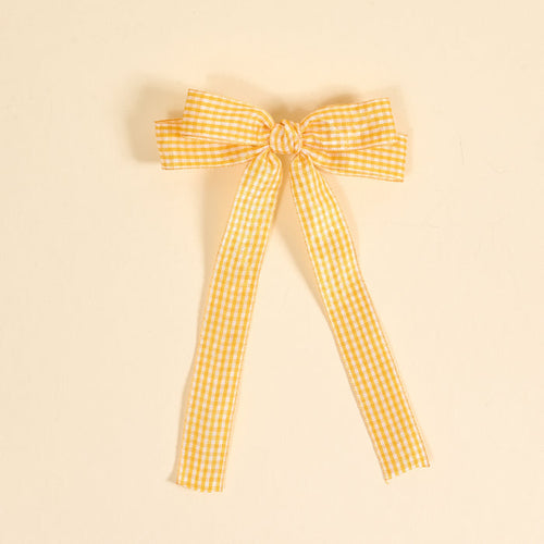 The Darling Effect's Gingham Yellow Double Take Hair Bow features yellow and white gingham ribbon tied into a bow with two long tails, set against a light beige background—perfect for hair accessories or as a stylish bow clip.