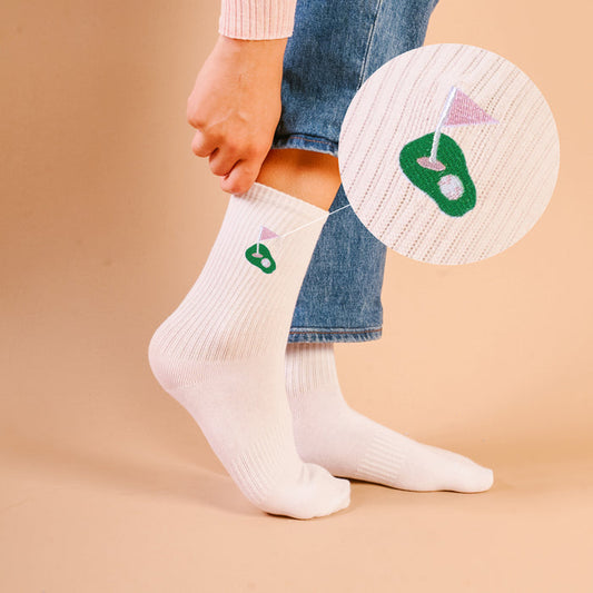 A woman pulls up The Darling Effect Golf Embroidered Crew Socks, featuring a green golf hole and purple flag design. A circular zoom on the beige background highlights the unique embroidered pattern of these stylish socks.