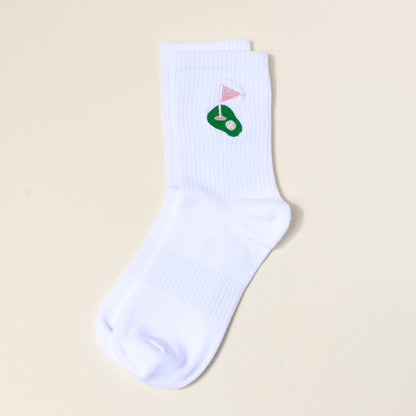 The Darling Effect Golf Embroidered Crew Socks: a white women's sock with a green putting green and pink flag design near the cuff, shown flat on a light beige background—ideal for fans of colorful, embroidered socks.