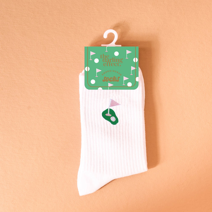 The Darling Effect Golf Embroidered Crew Socks, featuring a white ribbed design with a small green golf course and pink flag, are shown hanging from green packaging against a peach background.