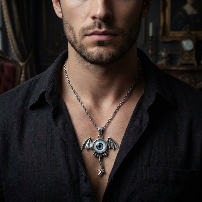 Man wearing a gothic silver necklace featuring a realistic eyeball pendant with bat wings and serpent tail design, displayed on an open black shirt against a dark vintage interior background