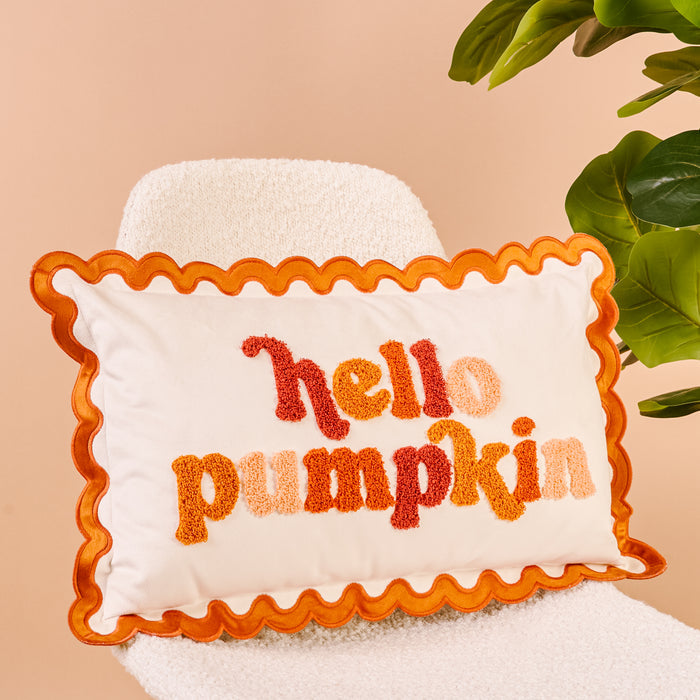 The Darling Effect's Hello Pumpkin Scalloped Rectangle Throw Pillow, featuring textured orange, red, and peach lettering on cream fabric, sits on a white boucle chair.