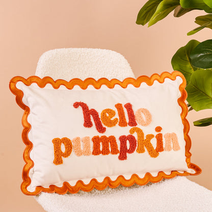The Darling Effect's Hello Pumpkin Scalloped Rectangle Throw Pillow, featuring textured orange, red, and peach lettering on cream fabric, sits on a white boucle chair.