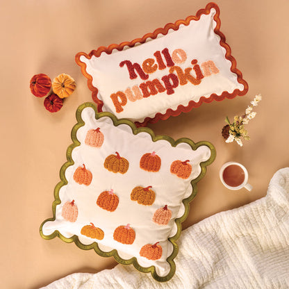 The Darling Effect's Hello Pumpkin Scalloped Rectangle Throw Pillow adds a cozy touch next to an embroidered pumpkin pillow, fabric pumpkins, tea, a small floral arrangement, and a white blanket on a beige surface.