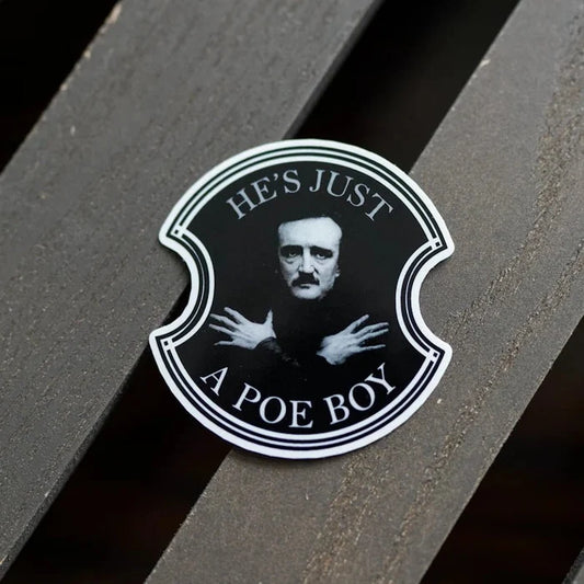 Black and white shield-shaped He's Just a Poe Boy sticker featuring Edgar Allan Poe portrait with arms crossed, placed on dark wood slat background — bookish gothic clear sticker by LitJoy Crate