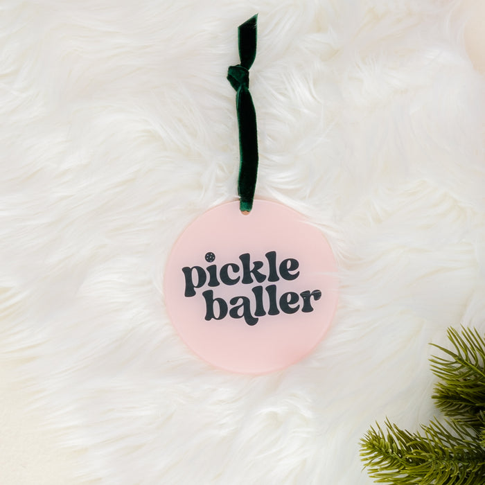 Add festive flair to your decor with the Holiday Tree Ornament - Pickleballer by The Darling Effect. This round pink acrylic ornament features the words “pickle baller” in bold black letters and hangs from a dark green ribbon; a touch of pine branch appears on the lower right, all set against a soft white furry background—making it an ideal choice for holiday celebrations.