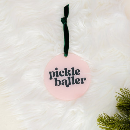 Add festive flair to your decor with the Holiday Tree Ornament - Pickleballer by The Darling Effect. This round pink acrylic ornament features the words “pickle baller” in bold black letters and hangs from a dark green ribbon; a touch of pine branch appears on the lower right, all set against a soft white furry background—making it an ideal choice for holiday celebrations.