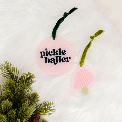 Two pink Holiday Tree Ornaments - Pickleballer by The Darling Effect, each adorned with a green velvet ribbon, rest on white faux fur. One ornament is round and features “pickle baller” in black lettering, while the other is shaped like a pickleball paddle. Ideal for festive holiday decor, green pine branches peek into the frame from the lower left corner.