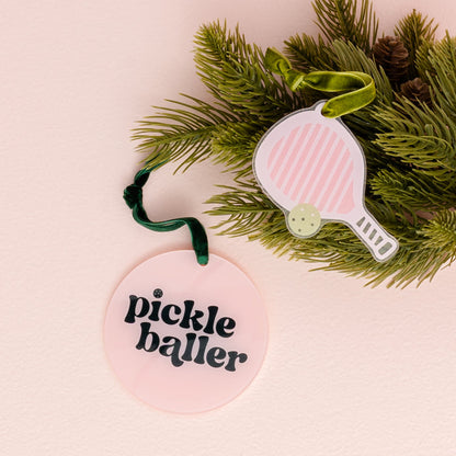 Two festive decorations hang from pine branches: one is The Darling Effect’s Holiday Tree Ornament - Pickleballer, a round pink acrylic ornament featuring “pickle baller” in bold black text, and the other is shaped like a pink-striped pickleball paddle—perfect touches for the holiday season.