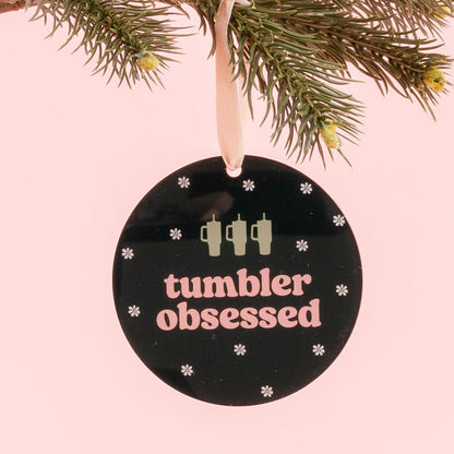 The Darling Effect’s Holiday Tree Ornament - Tumbler Obsessed is a round black acrylic ornament featuring the phrase “tumbler obsessed,” three tumbler icons, and delicate snowflakes. Hanging from a pine branch against a pink background, it’s the perfect festive addition to your holiday décor.