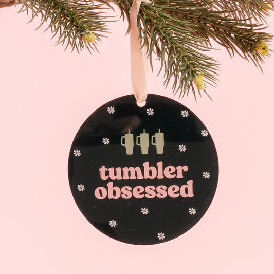 The Darling Effect’s Holiday Tree Ornament - Tumbler Obsessed is a round black acrylic ornament featuring the phrase “tumbler obsessed,” three tumbler icons, and delicate snowflakes. Hanging from a pine branch against a pink background, it’s the perfect festive addition to your holiday décor.
