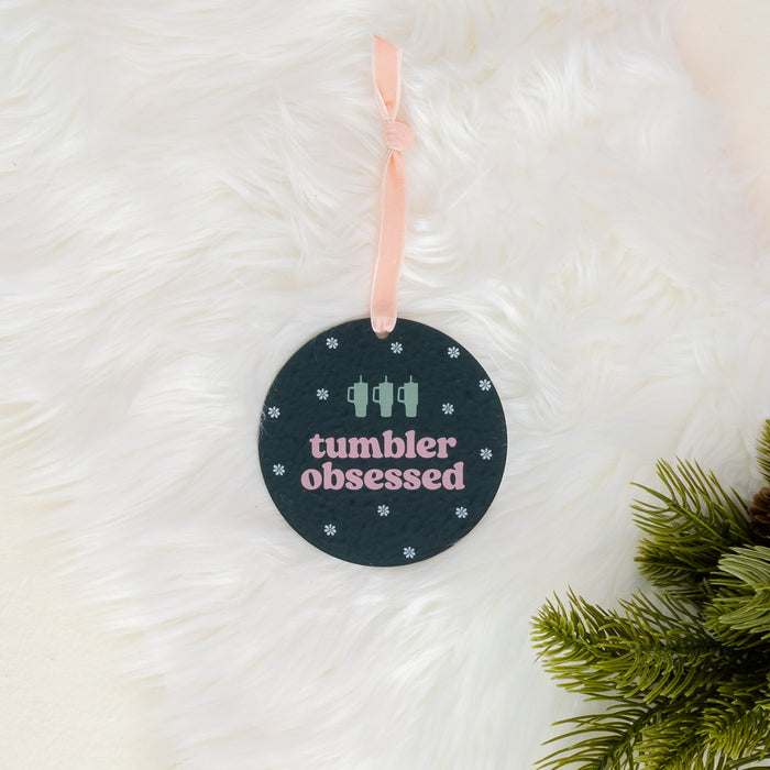 The Darling Effect’s Holiday Tree Ornament – Tumbler Obsessed features a round acrylic design with “tumbler obsessed” in pastel letters, two cup icons, and small snowflakes. Hanging from a pink ribbon on a white furry background with green pine branches, this ornament makes the perfect festive decoration for the holiday season.