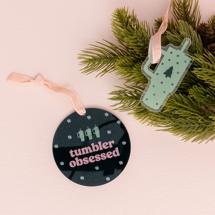 The Darling Effect’s Holiday Tree Ornament - Tumbler Obsessed features a round black acrylic design with “tumbler obsessed” written below three tumblers, as well as a green cup-shaped ornament adorned with a tree and hung from pine branches. Both festive decorations are finished with pink ribbon ties, bringing charming holiday cheer to your décor.