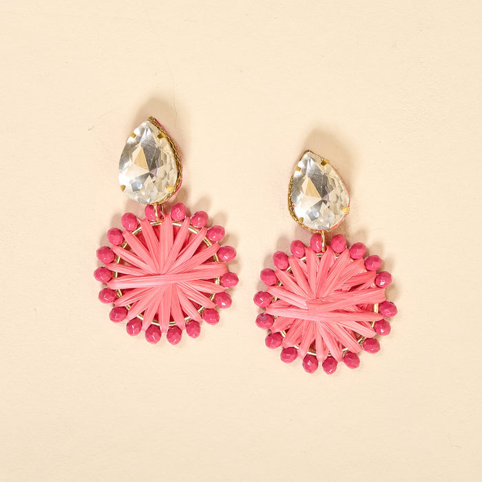 The Darling Effect's Hot Pink Radiant Gem Earrings are lightweight, boho-style earrings with clear teardrop gems atop round pink woven designs accented by small pink beads. Set against a light beige background.