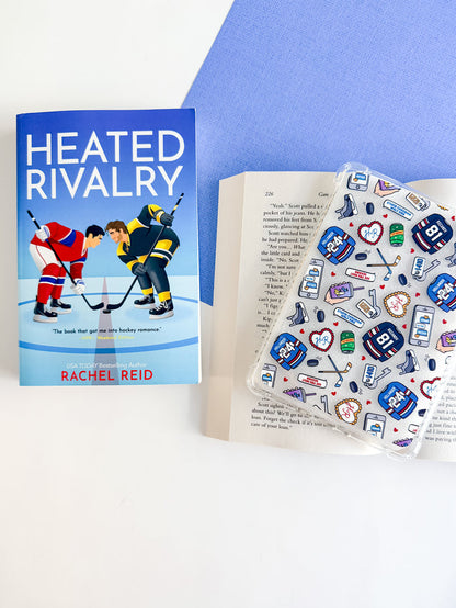 Book titled 'Heated Rivalry' by Rachel Reid with a clear case containing stickers on a white surface.