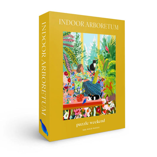 The Puzzle Weekend Indoor Arboretum 500 Piece Jigsaw Puzzle comes in a yellow box and features an illustration of a person and two cats amid lush houseplants.