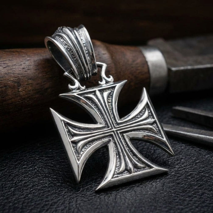 Sterling silver Iron Cross pendant with ornate detailing and decorative bail, displayed on black leather surface with wooden handle knife in background, featuring vintage Gothic style design with concave arms and textured border accents