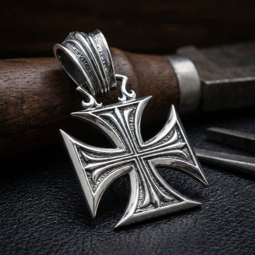 Sterling silver Iron Cross pendant with ornate detailing and decorative bail, displayed on black leather surface with wooden handle knife in background, featuring vintage Gothic style design with concave arms and textured border accents