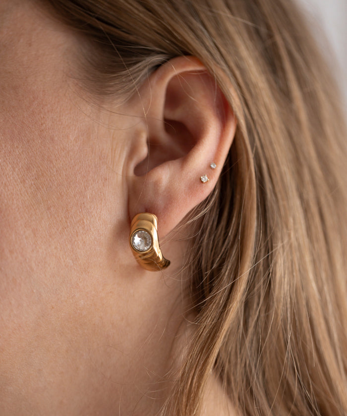 Close-up of a person wearing WALD Berlin's Isla Bonita Ready or Not earring, featuring a gold design with a clear Swarovski stone, complemented by two small studs on their upper ear, exuding an 80s vibe.
