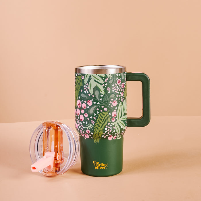 The Darling Effect’s Jolly Sprig 20 oz Lifestyle Flip Straw Tumbler features triple-walled insulation, a large handle, and a green floral-and-leaf design. The clear lid with a pink straw sits next to the mug on a beige background.