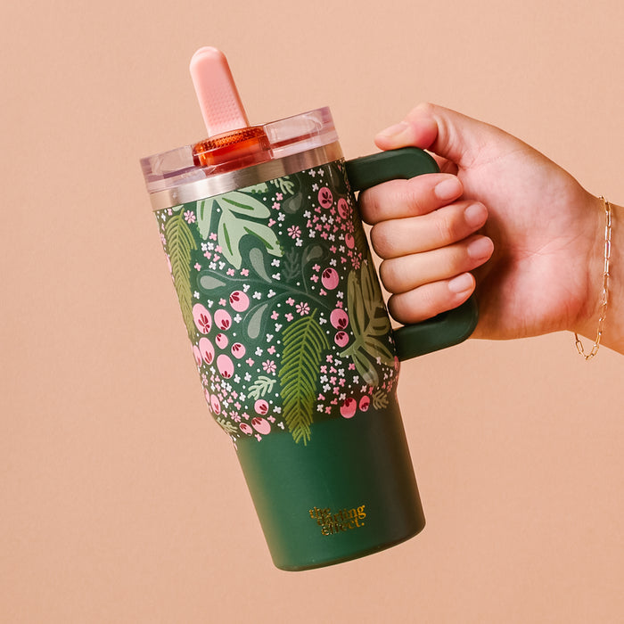 A hand holds The Darling Effect Jolly Sprig 20 oz Lifestyle Flip Straw Tumbler, featuring triple-walled insulation, a pink straw, and a dark green floral design with pink berries and white flowers.