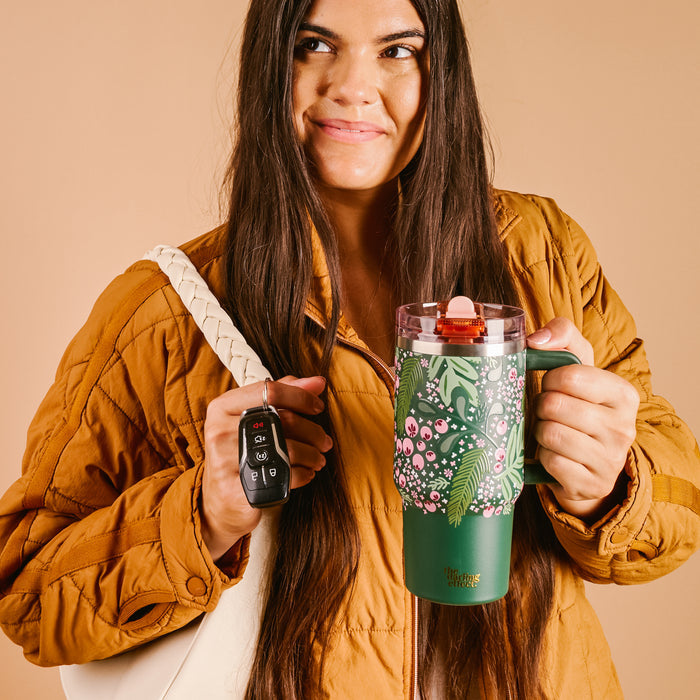 A woman with long brown hair in a tan jacket smiles while holding The Darling Effect Jolly Sprig 20 oz Lifestyle Flip Straw Tumbler and car keys, along with a white woven bag, against a beige background.