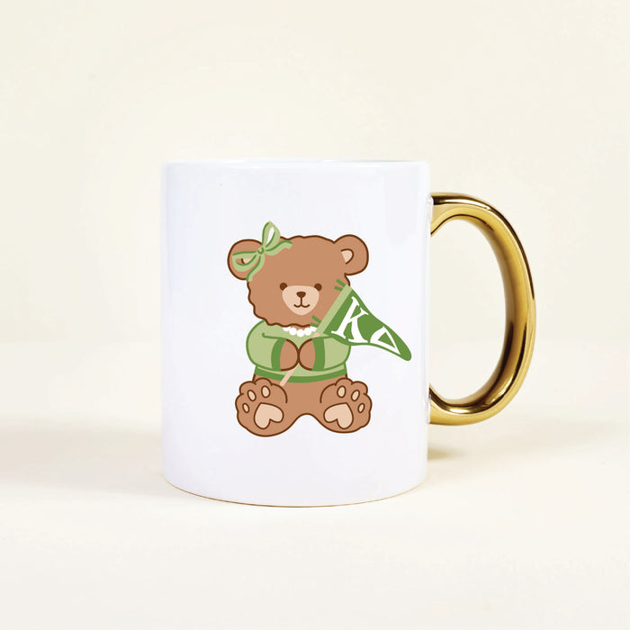 The Darling Effect’s Kappa Delta 11 oz Cozy Cute Ceramic Mug features a white base, gold handle, and an adorable teddy bear in a green KA headband, scarf, and pearls—a perfect addition to your collection.