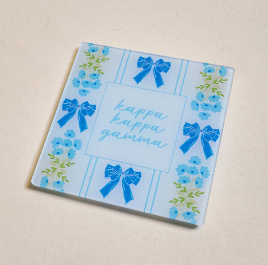 The Darling Effect’s Kappa Kappa Gamma Acrylic Coaster is a square blue piece with ribbon and floral accents, featuring "kappa kappa gamma" in light blue cursive—perfect for showing chapter pride or gifting your Big/Little.