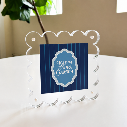 The Kappa Kappa Gamma Square Scalloped Acrylic Frame by The Darling Effect features a blue striped center with the sorority name in white, shown standing on a white table near a green plant and window.