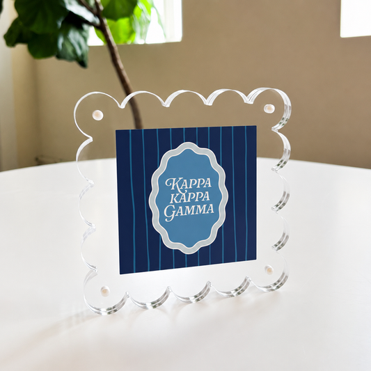 The Kappa Kappa Gamma Square Scalloped Acrylic Frame by The Darling Effect features a blue striped center with the sorority name in white, shown standing on a white table near a green plant and window.