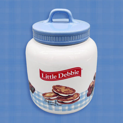 A Little Debbie gingham cookie jar with a white body and a light blue lid featuring a handle. The jar is decorated with a light blue gingham border near the bottom and showcases an illustration of Oatmeal Creme Pies labeled in cursive. Above the design, the red Little Debbie logo is prominently displayed.