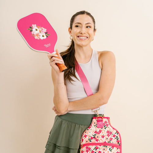 A smiling woman in a gray tank top and green skirt holds The Darling Effect's Lively Flora Pink Pickleball Paddle with a pink floral design and carries a matching floral bag, standing against a neutral background.
