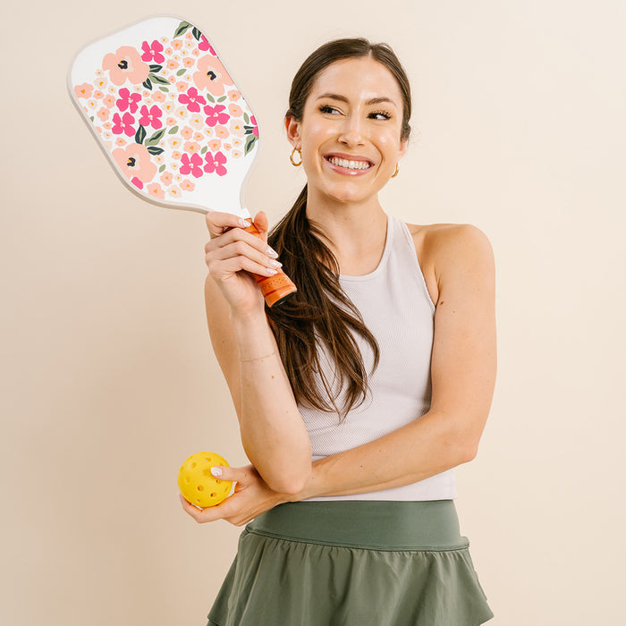 A woman in athletic wear smiles as she holds The Darling Effect’s Lively Flora Pink Pickleball Paddle and a yellow pickleball, proudly displaying her love for premium paddles.