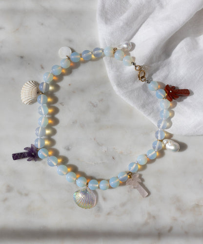 The Mexicana La Bamba Necklace by WALD Berlin features handmade iridescent opalite beads, shell, pearl, and stone cross charms—jewelry inspired by Mexicana style and displayed on marble with a white cloth.