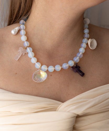 Wearing the WALD Berlin Mexicana La Bamba Necklace, which features opalite beads and palm tree, cross, shell, and seashell pendants, paired with an off-shoulder cream top.