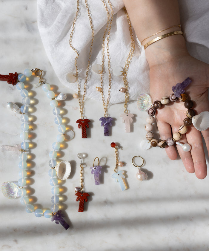 A hand with gold bracelets displays WALD Berlin handmade jewelry, including the Mexicana La Bamba Necklace with opalite beads, plus bracelets, earrings, and keychains featuring cross, shell, and palm tree pendants on a marble surface.