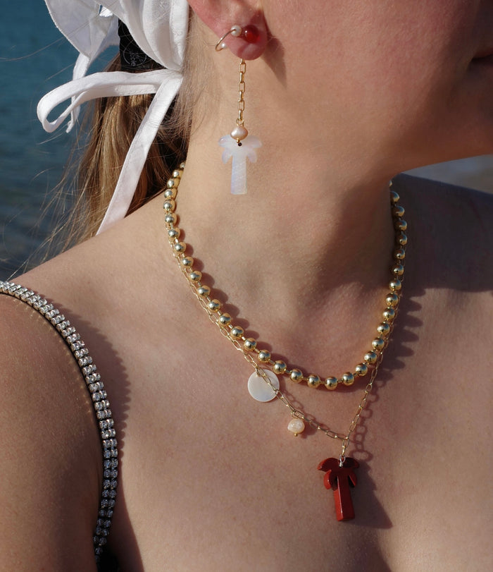 A woman stands near water wearing a white headscarf, layered gold necklaces with pendants, and WALD Berlin's Mexicana Mariachi Earrings Pearl.