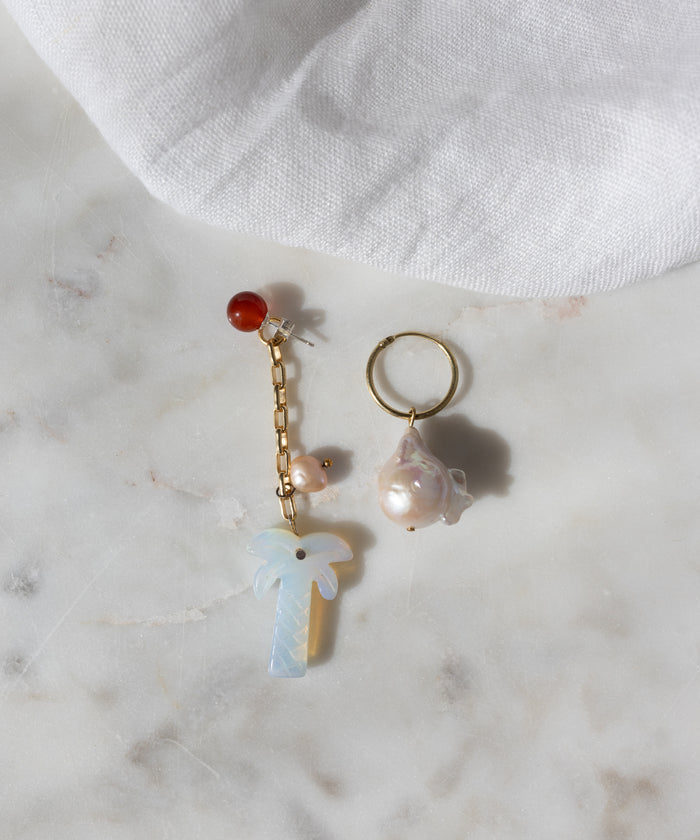 The Mexicana Mariachi Earrings Pearl by WALD Berlin are mismatched gold earrings with pearls, red bead, chain, and palm tree charm, displayed on marble. Handmade in Germany. A piece of white fabric is visible in the corner.