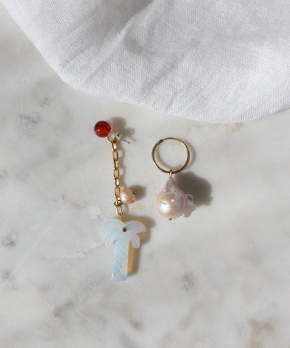 The Mexicana Mariachi Earrings Pearl by WALD Berlin are mismatched gold earrings with pearls, red bead, chain, and palm tree charm, displayed on marble. Handmade in Germany. A piece of white fabric is visible in the corner.
