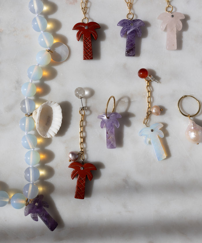 WALD Berlin’s Mexicana Mariachi Earrings Pearl and other palm tree necklaces, colorful earrings, a pearl ring, and a seashell are displayed on marble with handmade asymmetric gold earrings from Germany.