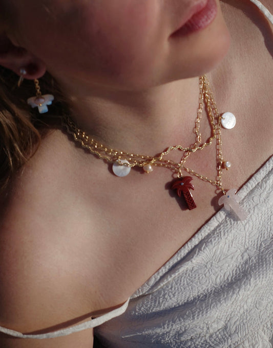 A person wearing layered gold necklaces, including WALD Berlin’s Mexicana Solo Amore Necklace with a hand-carved stone palm pendant and matching earrings, dressed in a white textured top, photographed from the shoulders up.