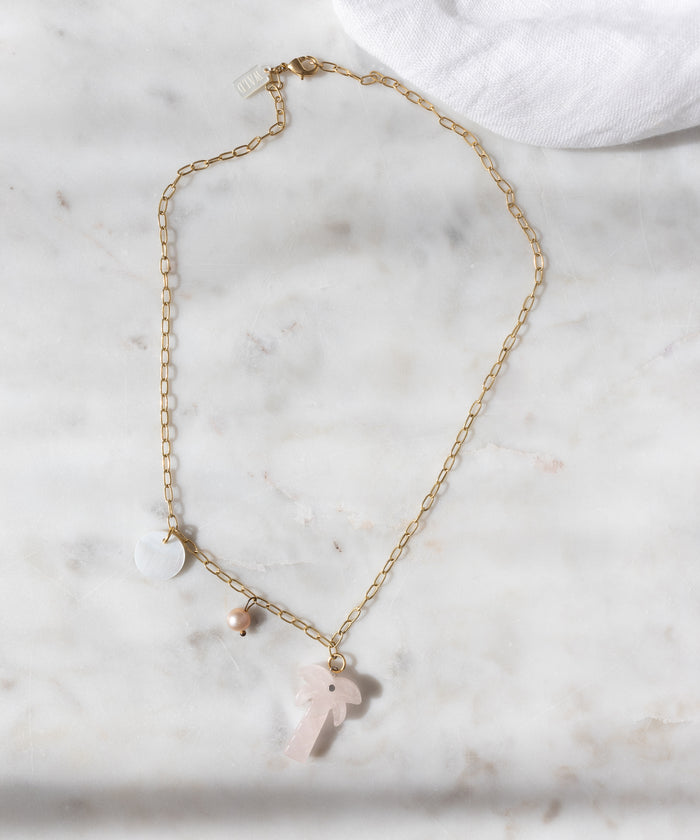 The WALD Berlin Mexicana Solo Amore Necklace is a handmade gold-plated piece with three pendants—a white disc, small bead, and light pink hand-carved stone palm—shown elegantly on marble.